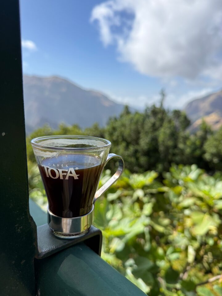 A glass of Cafés TOFA coffee resting on the terrace railing, mountains behind