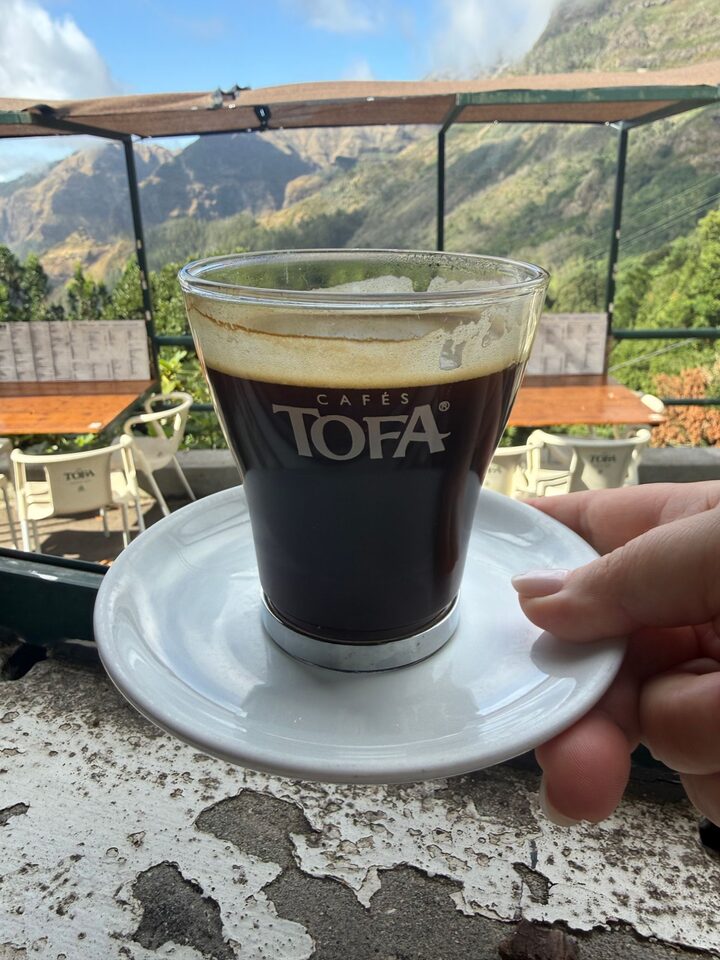 A glass of Cafés TOFA coffee held up in front of the terrace and the valley
